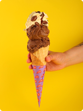 Una mano sostiene un cono de gofre con bolas de helado de chocolate y vainilla, cubierto de trocitos de chocolate, sobre un fondo amarillo brillante. El cucurucho está envuelto en un papel morado con motivos rojos.